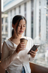 A beautiful Asian woman using her smartphone and holding a coffee cup while standing by the window.