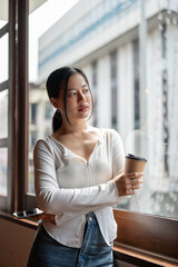 A beautiful and thoughtful Asian woman stands by the window with a coffee cup in her hand.