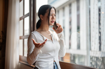 An angry and dissatisfied Asian woman is talking on the phone while standing by the window. - obrazy, fototapety, plakaty