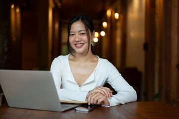 An attractive Asian woman is smiling at the camera and sitting at a table in a cafe with her laptop.
