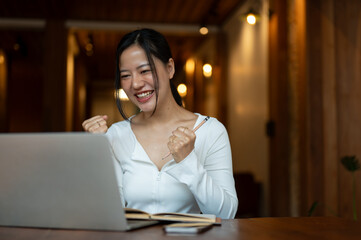 A cheerful woman is looking at her laptop screen with a happy and excited face, showing her fists.