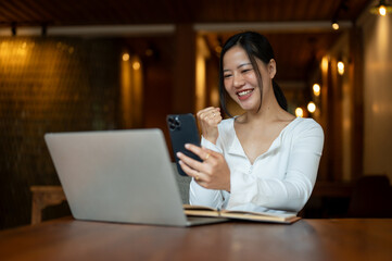 A cheerful Asian woman is looking at her smartphone screen with a happy and excited face
