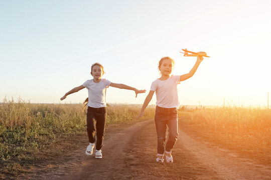 Joyful sisters run along road in field playing with toy plane. Concept of entertainment in childhood and imitation of flight. Imagination and play