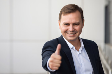 Confident adult businessman in formal suit stands near office and smiles raising thumb-up. Mature office worker expresses successful deal end, copyspace