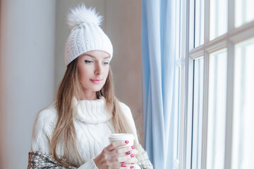 Portrait of pretty model woman brunette wearing season casual clothes white sweater and knitted hat  holding hot dreank in her hand posing at cozy home