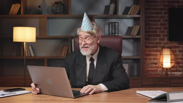 Happy aged business manager in paper cone hat receiving congratulations via video call app on laptop in office. Smiling professional expressing gratefulness for greetings and wishes from colleagues.