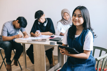 Young Asian student girl holding a digital tablet and smiling at the camera while classmates are studying in the background