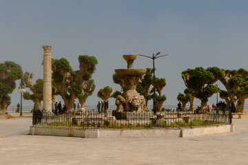 Cherchell, Algeria, 10 23 2023 : main square of Cherchell in a misty atmosphere. Algerian people, sand, plane trees, Roman column, fountain.
