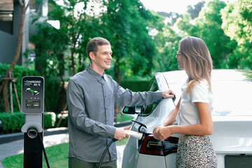 Young couple travel with EV electric car charging in green sustainable city outdoor garden in...