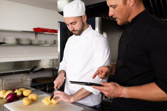 Caucasian male chef teacher in using tablet and guiding student in cutting potatoes in kitchen - Powered by Adobe