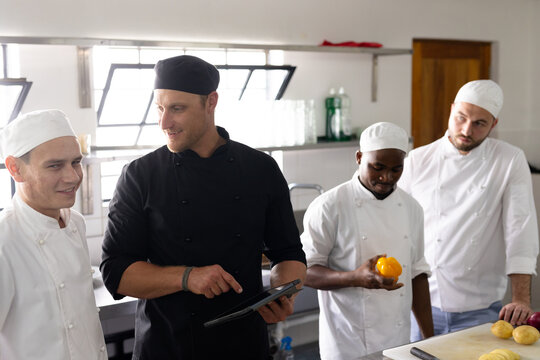 Diverse chef teacher using digital tablet and teaching cooking to male students in kitchen