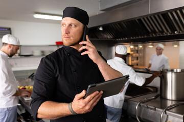 Diverse male chef teacher using tablet and talking over smartphone with students in background