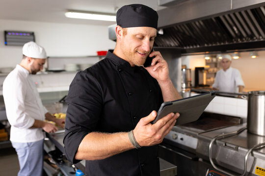 Caucasian male chef teacher using tablet and talking over cellphone with students in background