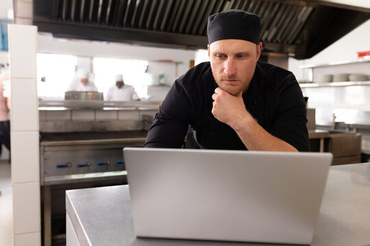 Serious caucasian male chef in black uniform thinking and working over laptop on counter in kitchen
