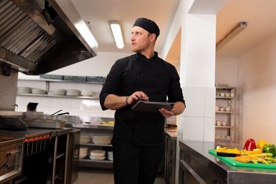 Caucasian male chef in black uniform holding tablet and looking away while standing in kitchen