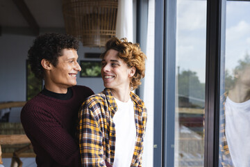 Happy diverse gay male couple embracing and looking at each other at window at home