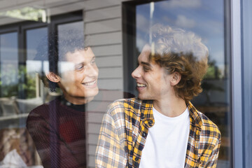 Happy diverse gay male couple embracing and looking at each other at window at home