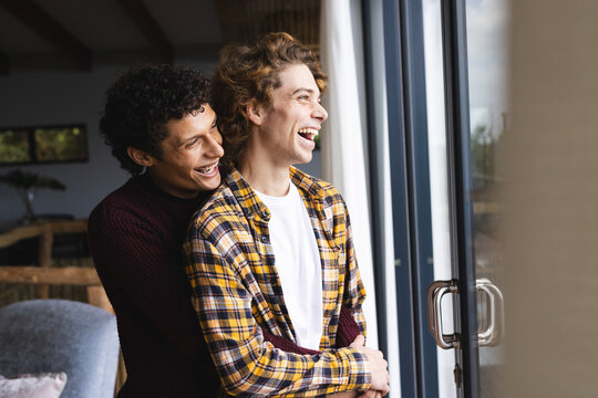 Happy diverse gay male couple embracing and looking through window at home