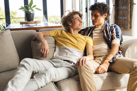 Happy diverse gay male couple sitting on couch and embracing at home