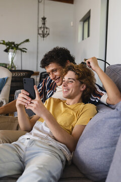 Happy diverse gay male couple using smartphone and embracing at home