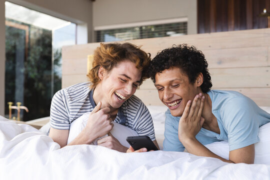 Happy diverse gay male couple lying on bed and using smartphone at home