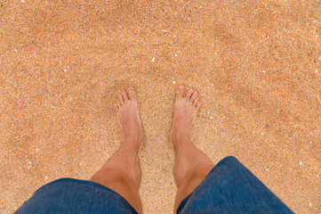 Fototapeta na wymiar Top view of male legs in blue shorts on golden sand in sunlight. Looking down at a man in shorts standing on the sea sandy beach