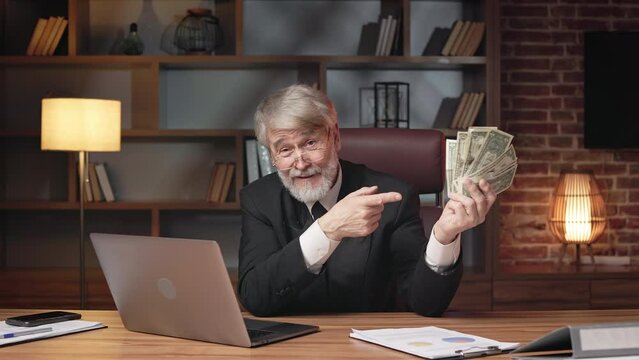 Positive older man in formal suit and glasses pointing at fan of paper money while staying in comfortable office. Professional business executive winking at camera while bragging about profit.