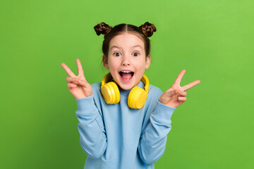 Photo of ecstatic funny schoolgirl with buns hairdo dressed blue pullover in headphones show v-sign...