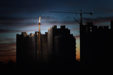 Tower cranes at new city building and apartment construction sites, red sunset sky and clouds, dark...