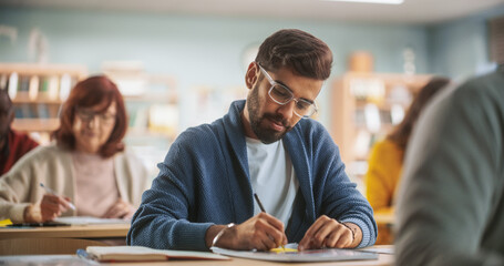 Portrait of an Indian Student Taking a Course in an International Adult Education Center. Young Man Wearing Glasses, Sitting Behind a Desk, Writing Down Notes Together with Diverse Colleagues - obrazy, fototapety, plakaty