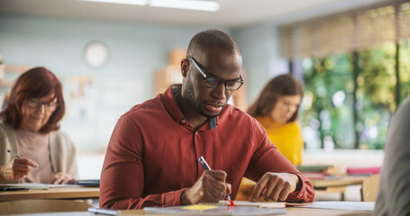 Handsome African Man Writing Down Notes in Notebook. Group of International Mature Students Studying in Classroom. Modern Adult Training Center Help People to Develop New Useful Skills Throughout Life - obrazy, fototapety, plakaty