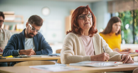 Portrait of Happy Senior Woman Taking a Course in an International Adult Education Center. Old Female Wearing Glasses, Sitting Behind a Desk, Using Textbook and Writing Down Notes in Notebook