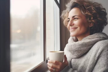 Poster Portrait of happy middle aged woman in cozy sweater holding a cup of hot drink and looking trough the window, enjoying the winter morning at home, side view © Jasmina