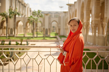 Charming Muslim woman in orange scarf, looking at camera, standing on the balcony against the mosque garden background