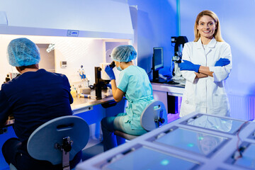 Beautiful successful mature blonde woman scientist wearing white coat and gloves in modern medical science laboratory with team of specialists on background.