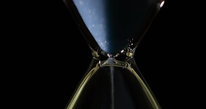 Pure blue fine sand flows inside traditional hourglass. Measure life expectancy on black background