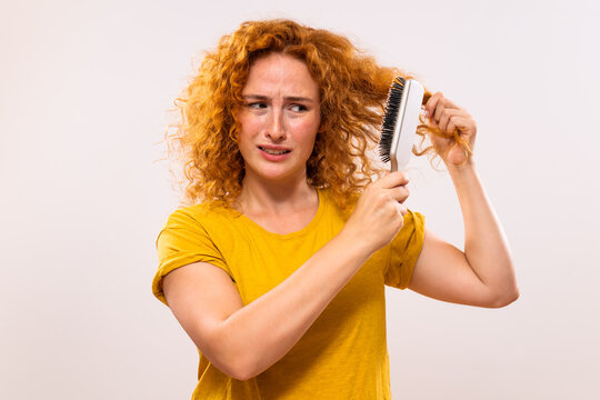 Angry ginger woman holding hairbrush and combing her curly hair.