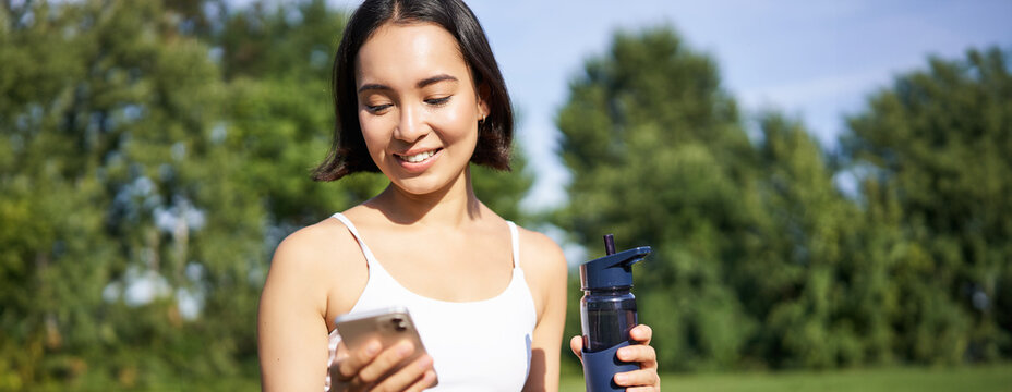 Smiling fitness girl drinks water, checks her app on smartphone and looks happy, stays hydrated on fresh air, sunny day in park