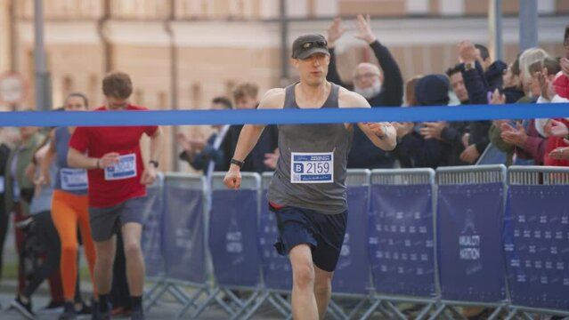 Slow Motion Portrait of Athletic Male Jogger Crossing the Finish Line in Marathon with the Audience Cheering. Happy Successful Man Celebrating Winning, Achieving his Goal at Finishing First in a Race