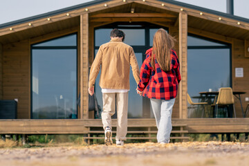 A guy and a girl walk together holding hands into wooden house with large panoramic windows