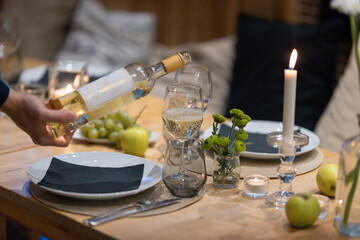 Man pours white wine into a glass - dining table set with candles and plates