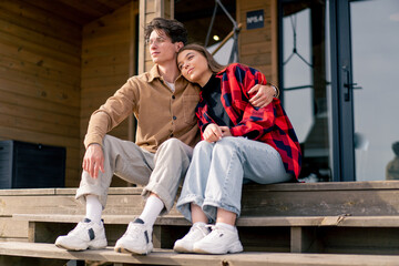 A young stylish couple in love is sitting together hugging on the steps of  large wooden cottage