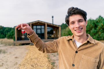 A guy holds a bunch of keys to his new house in the background of a wooden cabin in woods in the countryside - obrazy, fototapety, plakaty