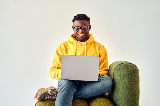 Sincere emotions and technology. Black male freelancer looking on laptop screen with cheerful smile while sitting on green lounge chair in studio. Young guy wearing googles and casual yellow hoodie.