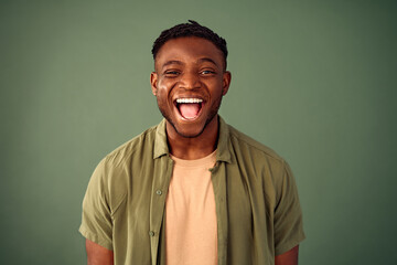 Frank facial expression. Portrait of young african american man smiling cheerfully with open mouth over green background. Handsome guy dressed in casual khaki shirt expressing happiness in studio. - obrazy, fototapety, plakaty