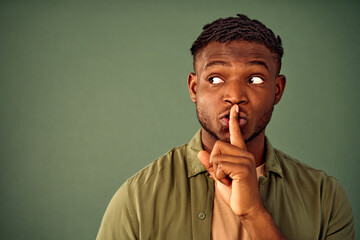 Keep silence, shh. Handsome african american dressed in unbuttoned khaki shirt holding forefinger near lips and looking aside while asking for silence. Isolated over green background. Copy space.