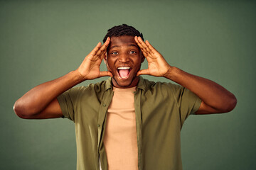 Surprise reaction. Amazed african man expressing astonishment with open mouth while standing over green background and looking at camera. Young guy grabbing head with two hands and making big eyes. - obrazy, fototapety, plakaty