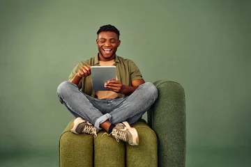 Zelfklevend Fotobehang Online leisure time. Happy black guy sitting in lotus pose on comfy design chair and scrolling social media on digital tablet over green studio background. Concept of youth and modern gadgets. © HBS