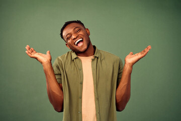 Oops, don't know. Pleasant african american guy shrugging shoulder and keeping palms wide open while standing over green background. Young man smiling with open mouth and expressing sincere emotions. - obrazy, fototapety, plakaty