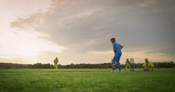 Professional Cricket Player in Blue Uniform and Protective Helmet and Pads Preparing to Hit the Ball and Win the Tournament. Two Young Amateur Teams Practicing on a Warm Evening at Sunset
